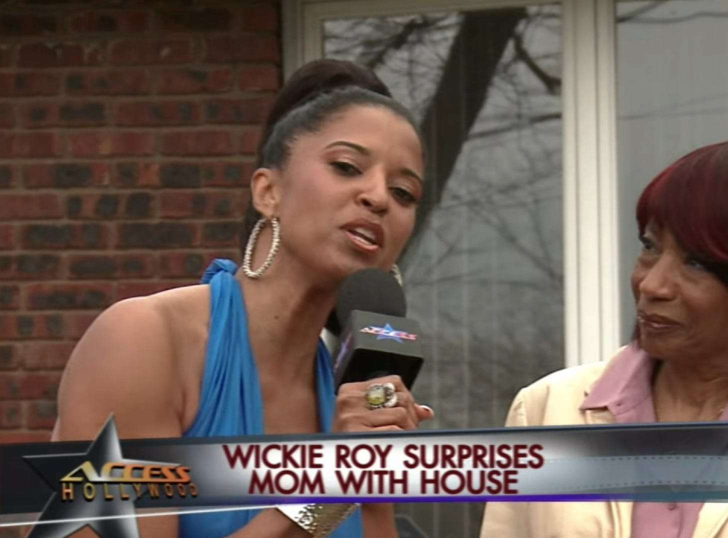 Renée Elise Goldsberry as Wickie Roy in a screenshot from Girls5eva Season 3: Wickie is on an Access Hollywood Segment, holding a microphone and addressing the camera. A chyron reads "Wickie Roy Surprises Mom With House"