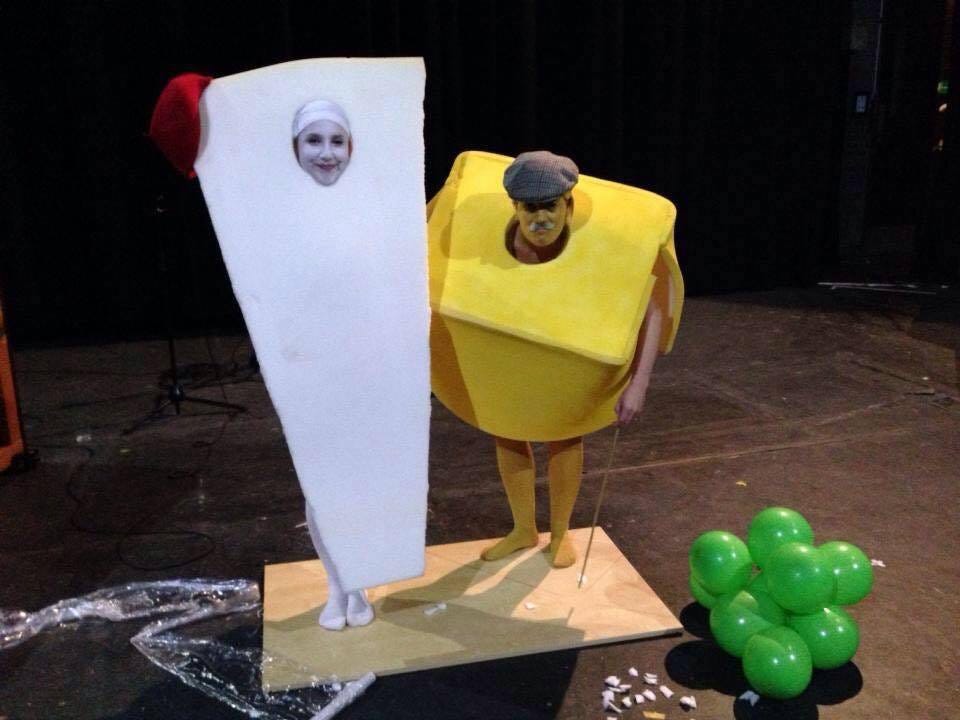 Two comedians dressed as pieces of cheese standing on a wooden board surrounded by green balloons to represent grapes