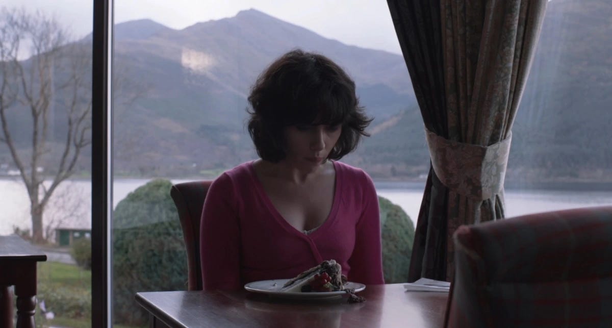 Scarlett Johansson as the alien in Under the Skin sits in a restaurant with a slice of cake in front of her.