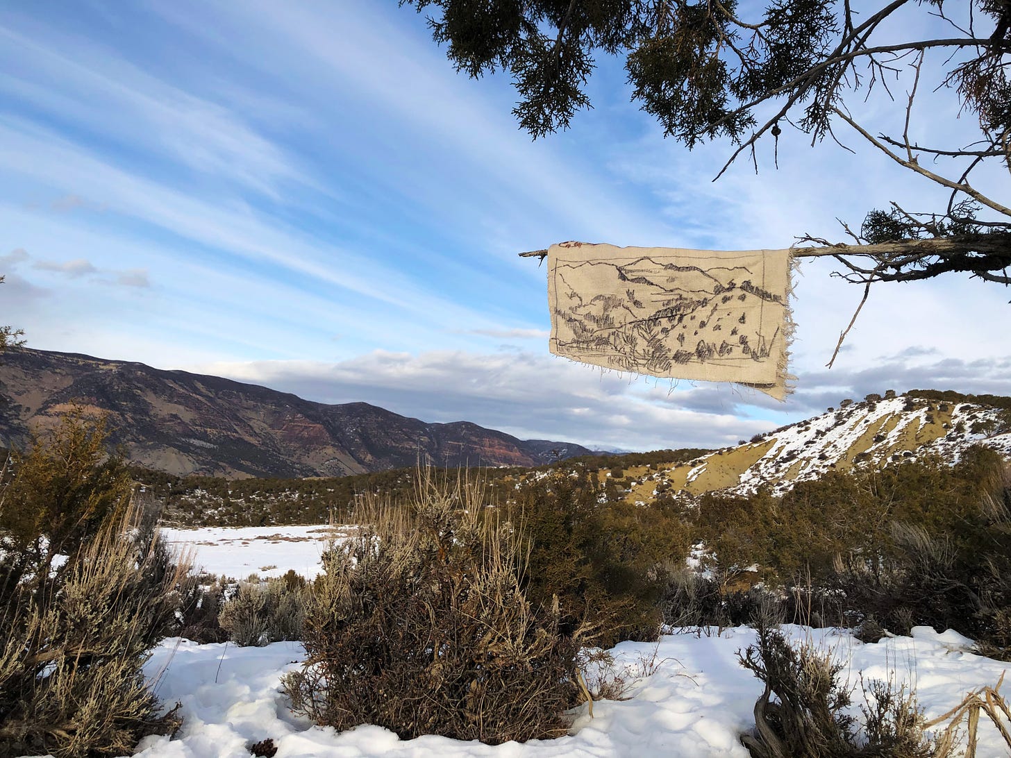 A winter landscape photograph. In the foreground is snow-covered scrub vegetation. The middle ground shows hills with patches of snow. In the background are mountains under a blue sky with wispy clouds. Hanging from a tree branch in the foreground is a piece of fabric with a hand-drawn landscape sketch on it.