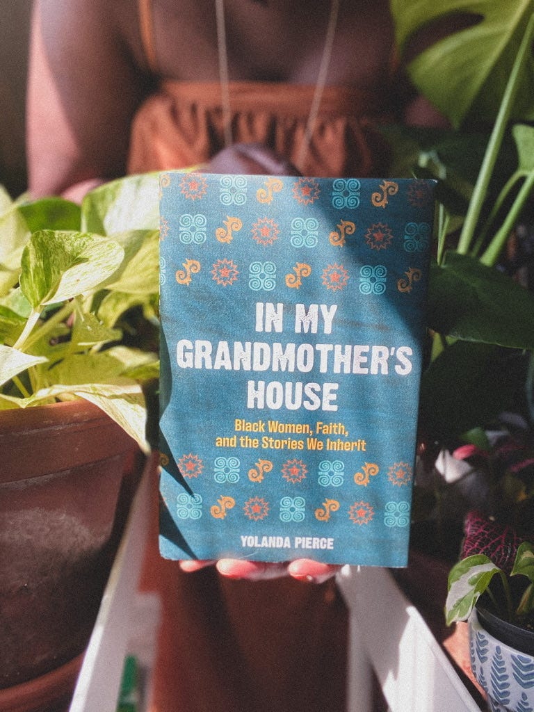 a picture of Rose holding up a copy of Yolanda Pierce's book "In My Grandmother's Hands." Rose ins blurred out in the background in a rustic organize dress with thin stripes. Light pours in from the left, and Rose and the books are flanked by house plants.