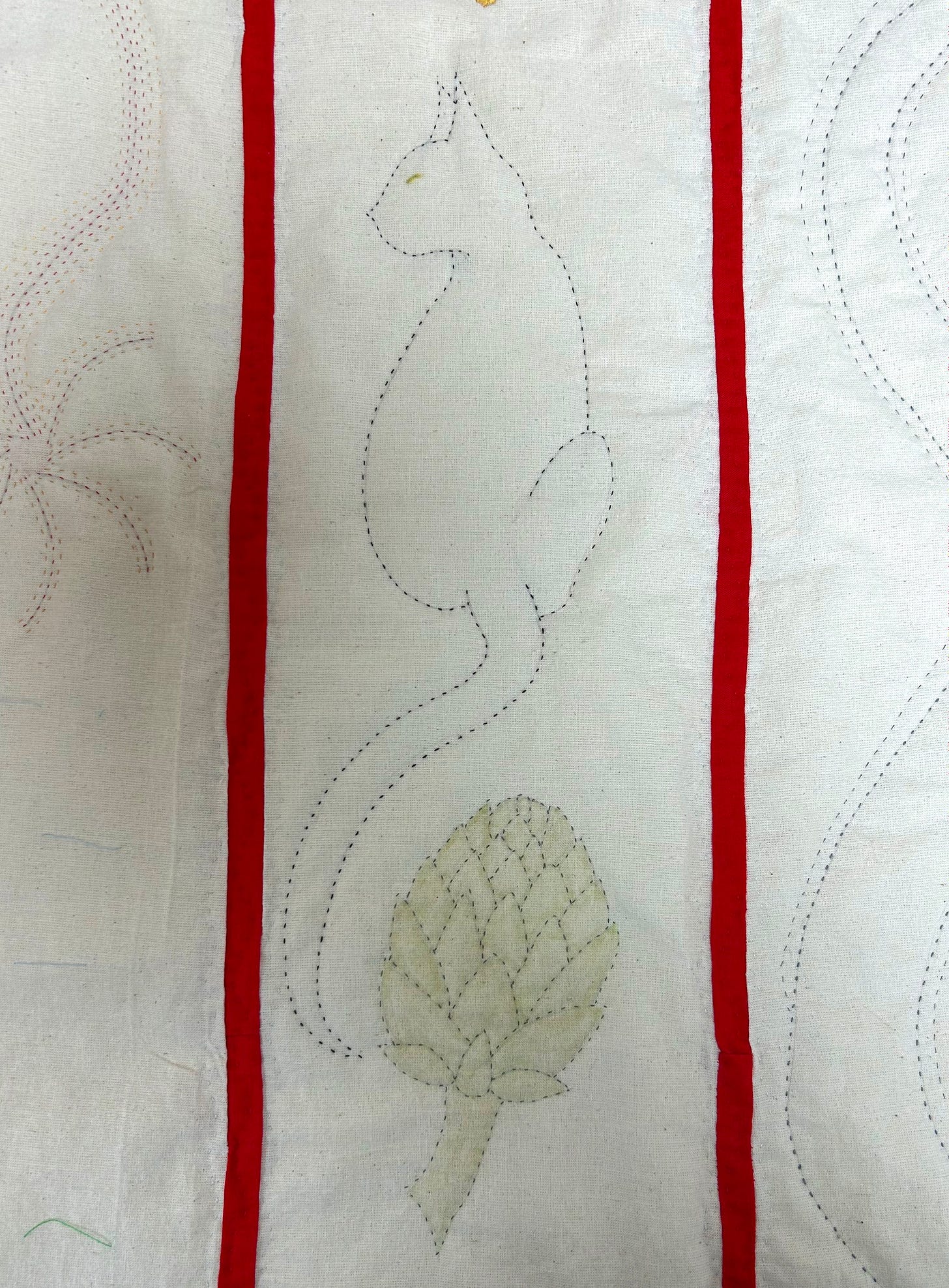 A stitched cat on cream coloured calico, sits above a stitched artichoke. There are two red strips on either side of the cat.