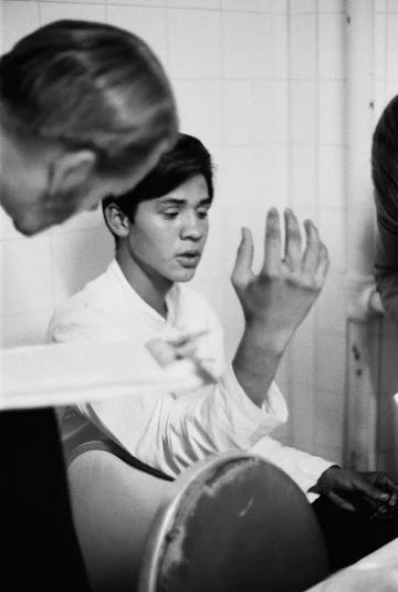 Black and white photo of a young Mexican man speaking to a white man. The white man is leaning in to listen to him. 