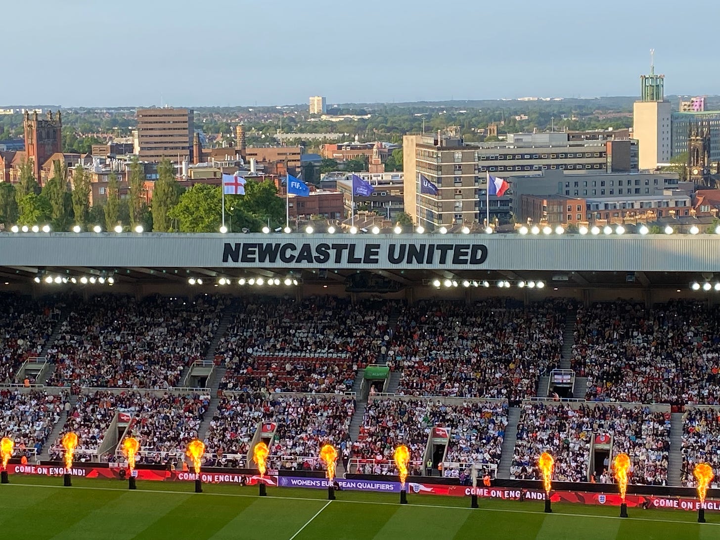 The view from the top of the Milburn Stand at Newcastle United. The pitch and some pyrotechnics in the foreground, the roof of the stand opposite and the view over the city in the sunlight behind that. 