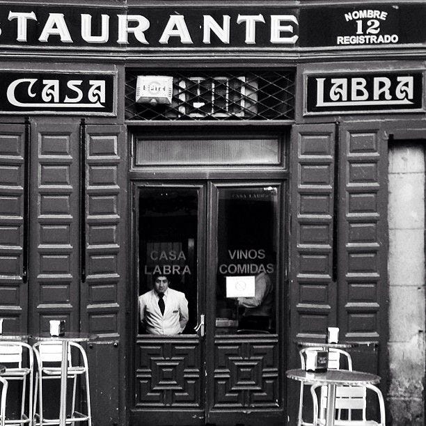 Black and white photo of the entrance to Casa Labra, a historic restaurant in Madrid, Spain. A man wearing a white jacket stands inside, looking out through the glass door. The signage above the door reads 'Casa Labra' and 'Restaurante.' Two high stools and a small table are visible outside the entrance.