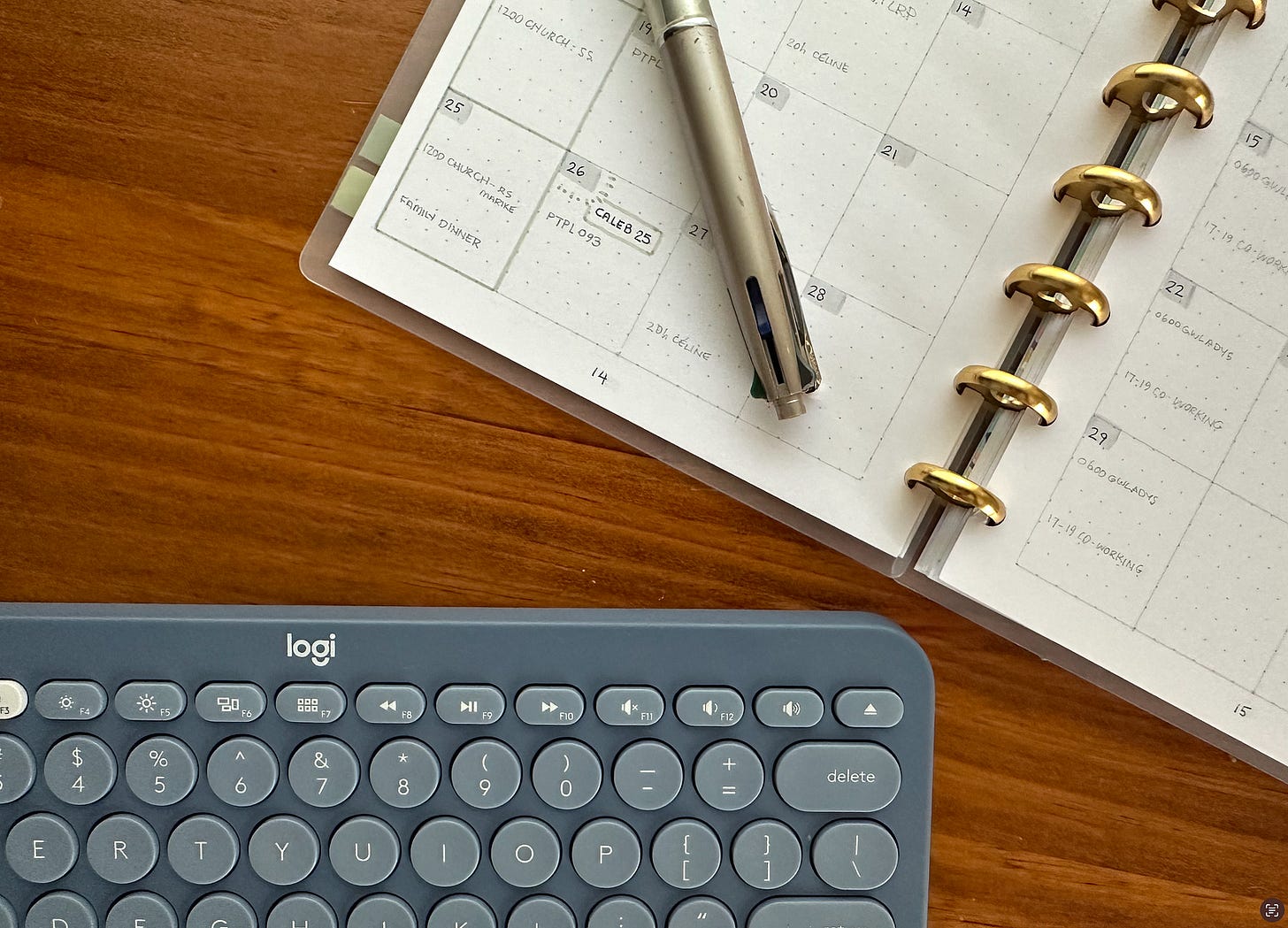 Blue-grey ‘logi’ keyboard and a discbound note book are sitting on a wooden table. We’re looking at a cropped view of them, from above. There’s a Pilot Dr Grip pen (missing its cap) sitting on the left hand planner page.