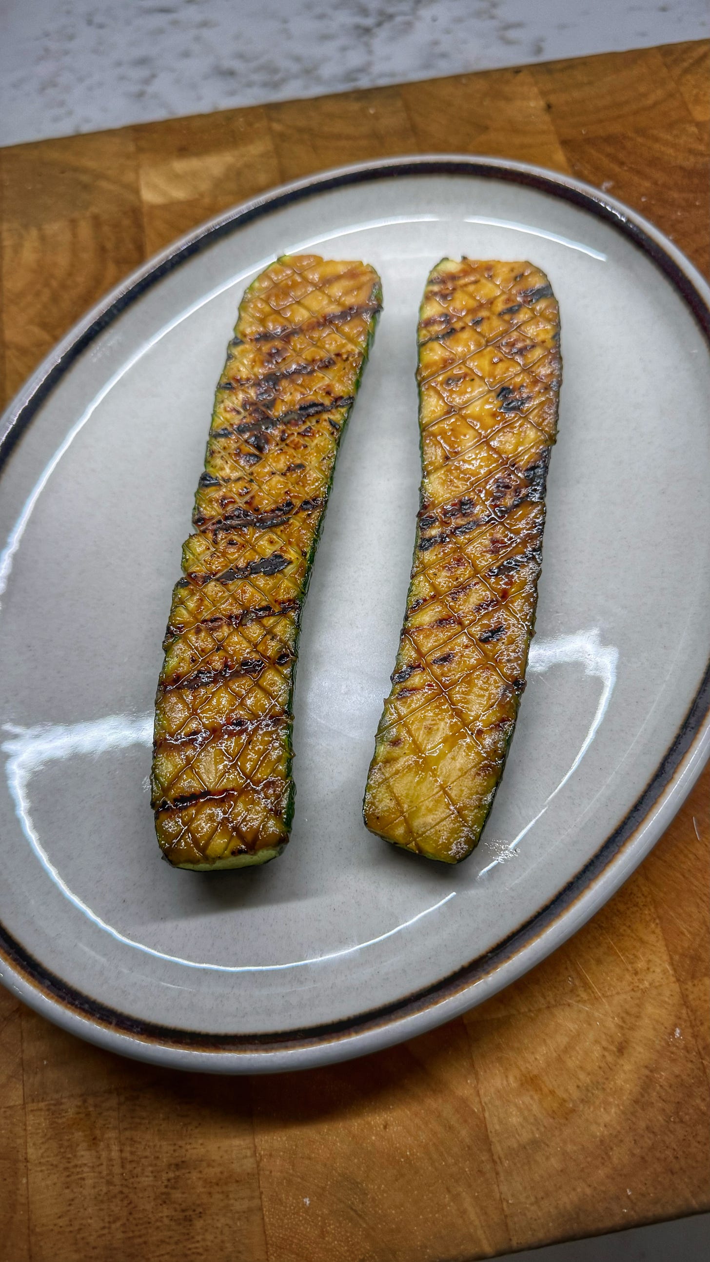 A serving dish with two large pieces of zucchini, halved lengthwise and having been scored in a criss-cross pattern, basted in sauce, and grilled until caramelized and grill marks are on the zucchini's flesh. 