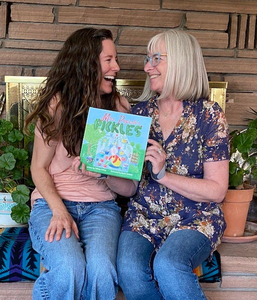 Two women authors sitting on a hearth facing each other and laughing, holding a book titled Mrs Popish's Pickles