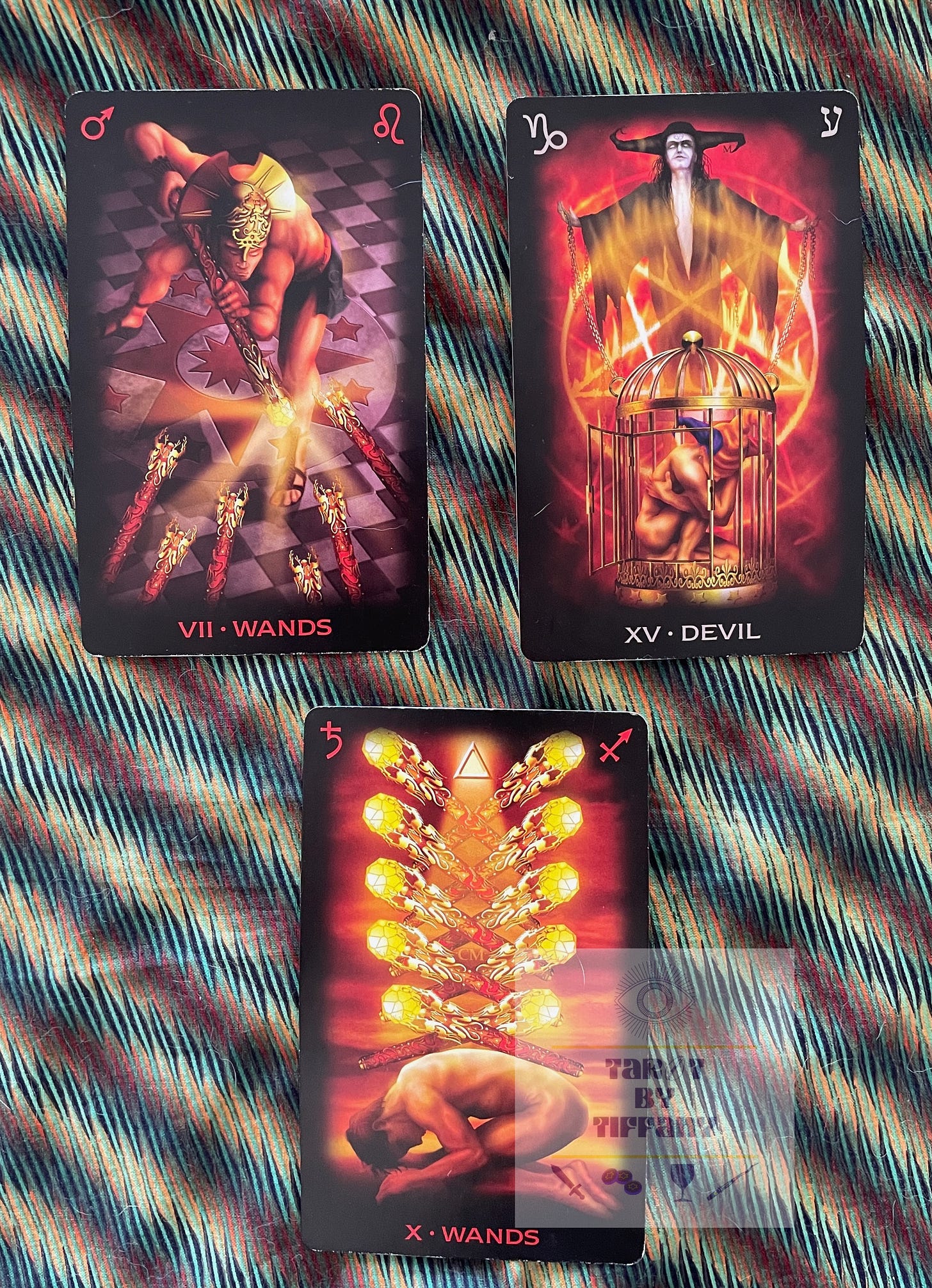 3-card spread using the Tarot of Dreams. Cards are on a cloth background with brown, dark green, and gold diagonal stripes. Top two cards (left to right): 7 of Wands and The Devil. Bottom card: 10 of Wands.