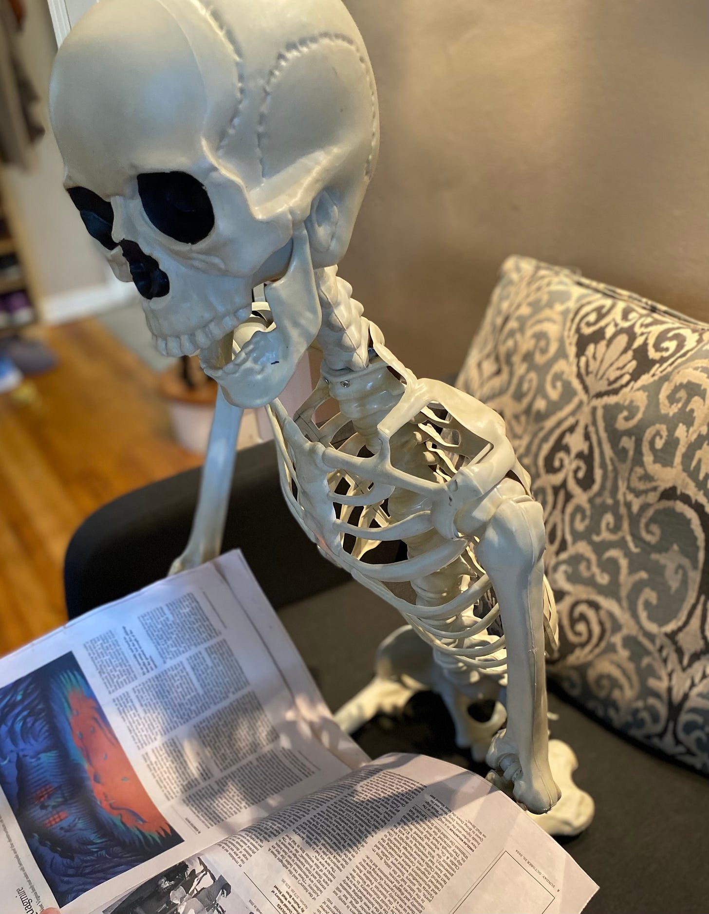 Plastic skeleton sitting on couch holding newspaper