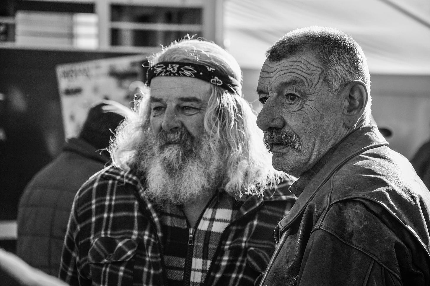 Two old men -one dressed in plaid wearing a beard and head band -the other hair cropped short .