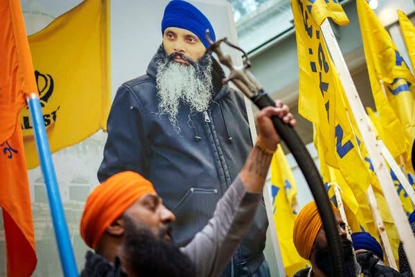 Men wearing turbans standing amid yellow flags and a poster of Hardeep Singh Nijjar, wearing a blue turban and a black hoodie. 