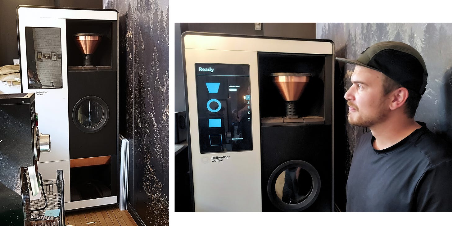 A six-foot tall electric roasting machine that looks like a Tesla vending machine is pushed against a wall featuring pine trees on the wallpaper. To the right, a coffee roasters looks off into the distance.