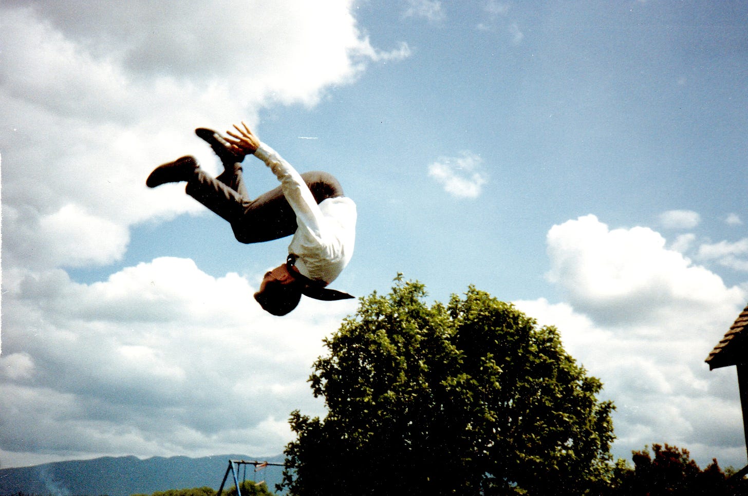 A Mormon missionary is captured against cloudy skies in a midair somersault, perhaps having bounced from a trampoline outside the frame.