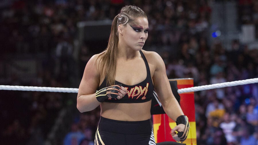 Image of Ronda Rousey in the middle of a wrestling arena wearing a black sports bra, black shorts, and black and orange gloves. Her hair is in a ponytail and she has dark eyeshadow on.