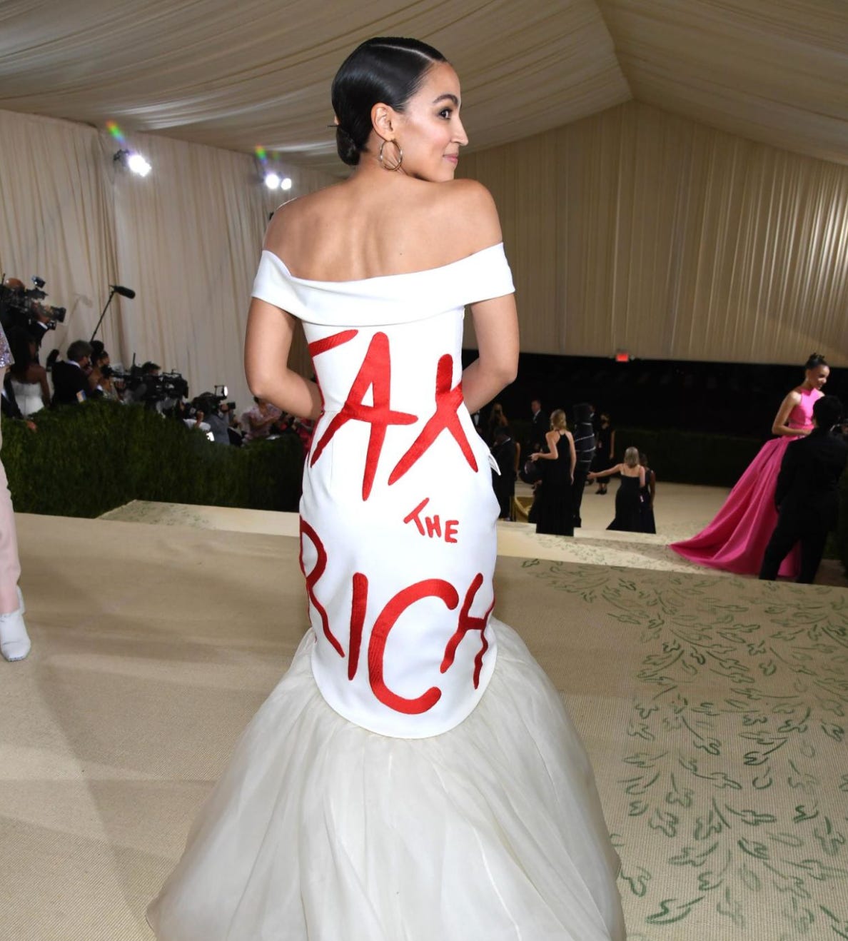 A light brown skinned woman (AOC) standing at the top of a staircase facing the stairs with her back to the camera. She has dark hair and is wearing a white figure hugging designer dress with “tax the rich” written on the back in shiny red font. Below is a crowd of richly dressed people in expensive suits and dresses.