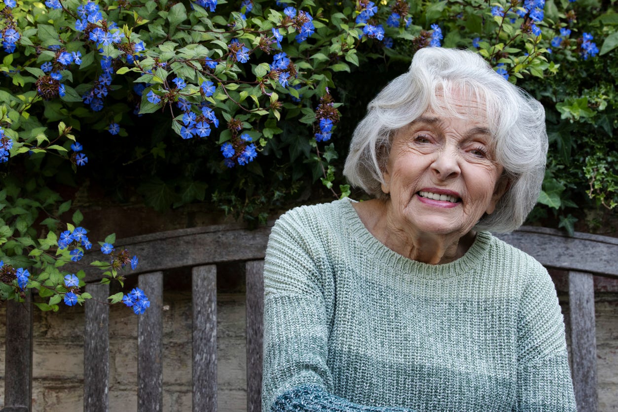 Smiling, older white woman with white hair, red lipstick and gray sweater on an outside deck with blue flowers in background.