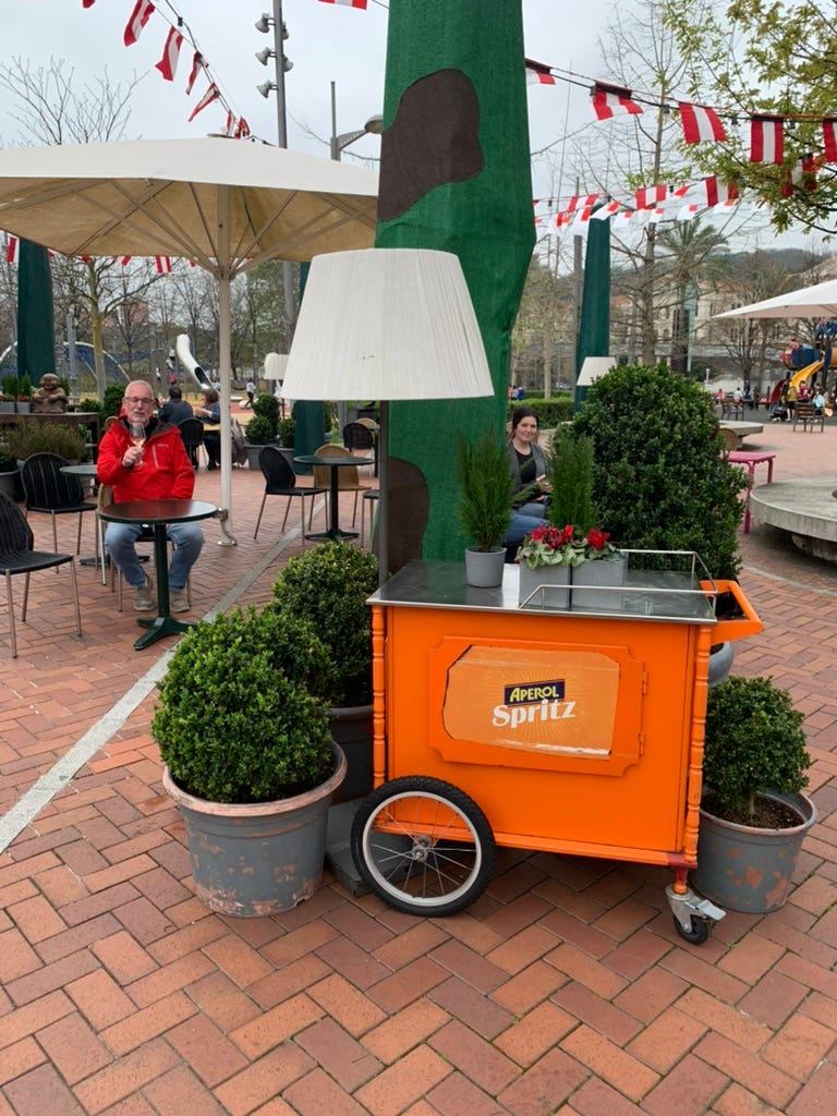 Jim enjoys a glass of wine at the outside bar in the park next to the museum. A bright orange cart complete with a floor lamp and flowers sits in the foreground.
