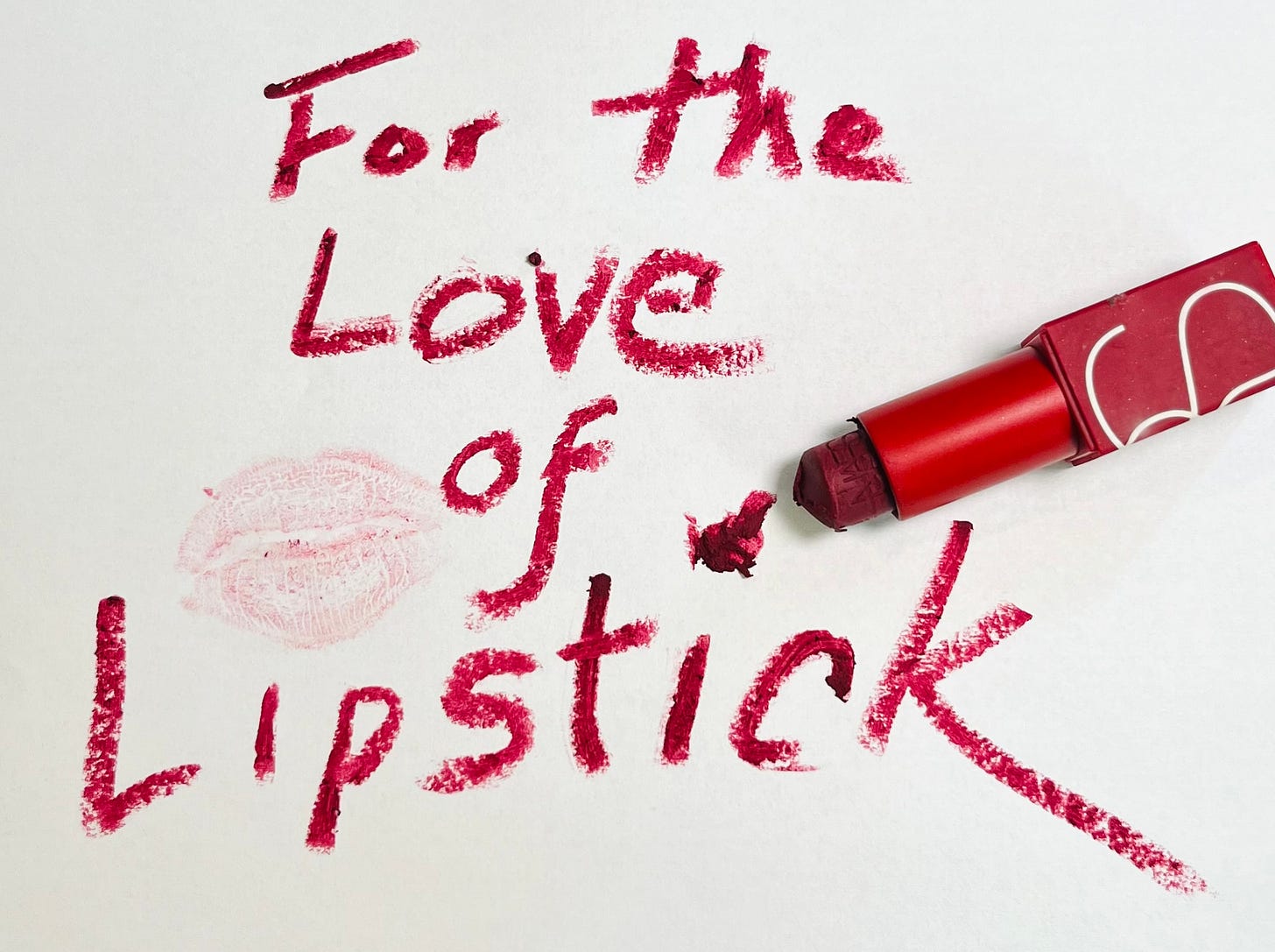 “For the Love of Lipstick” is written in lipstick on a piece of white paper. There is a lipstick kiss mark on the paper and the lipstick tube used to write the message rests on the paper as well. 