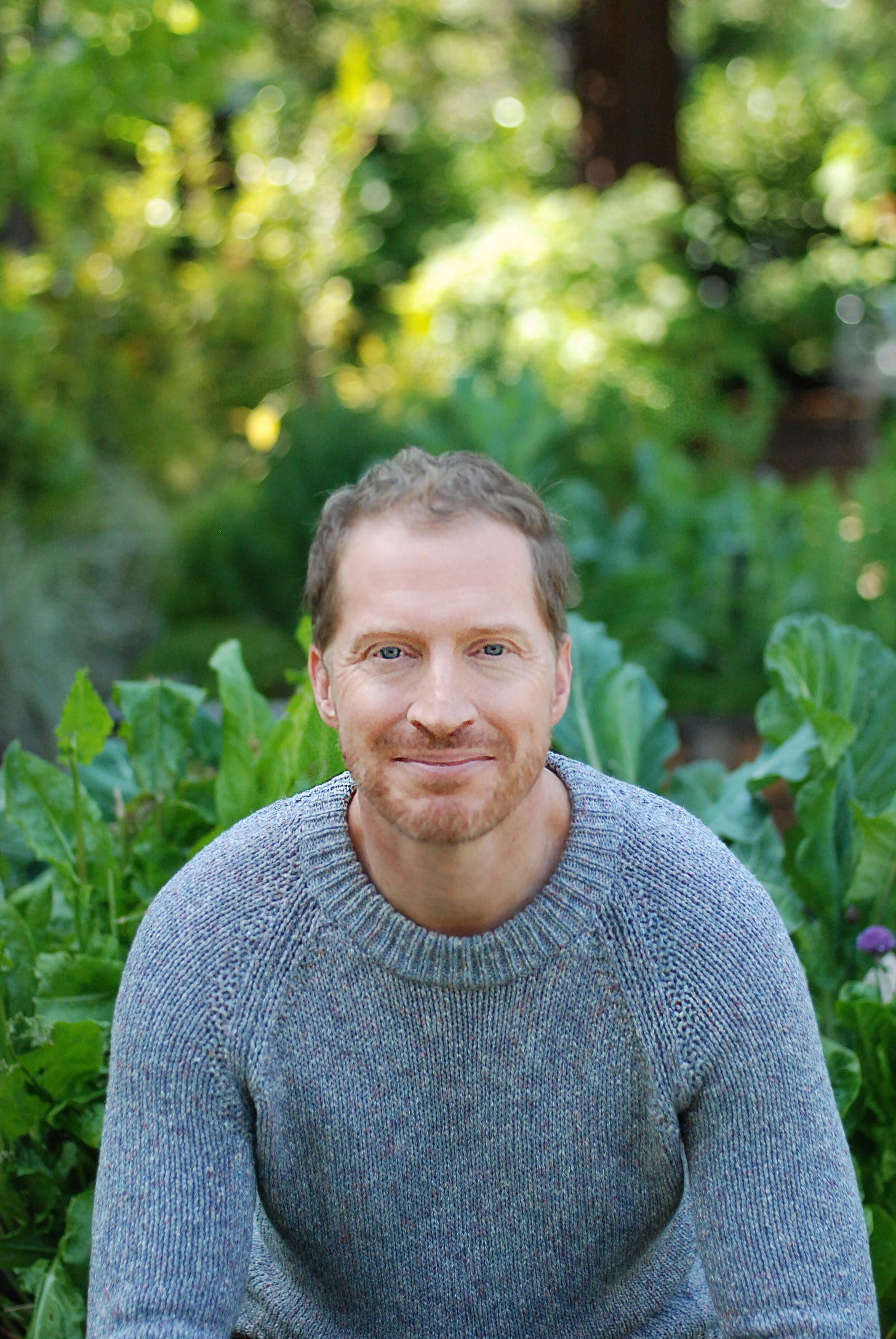 Author Andrew Sean Greer looking every one of his 51 years with sparse curly hair, a reddish beard and a winning smile, sitting before what seems to be a kale garden