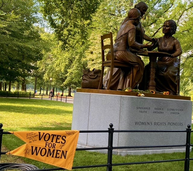 The Women's Rights Pioneers Monument from the side, where a visitor has affixed a yellow Votes for Women pennant to the fence.