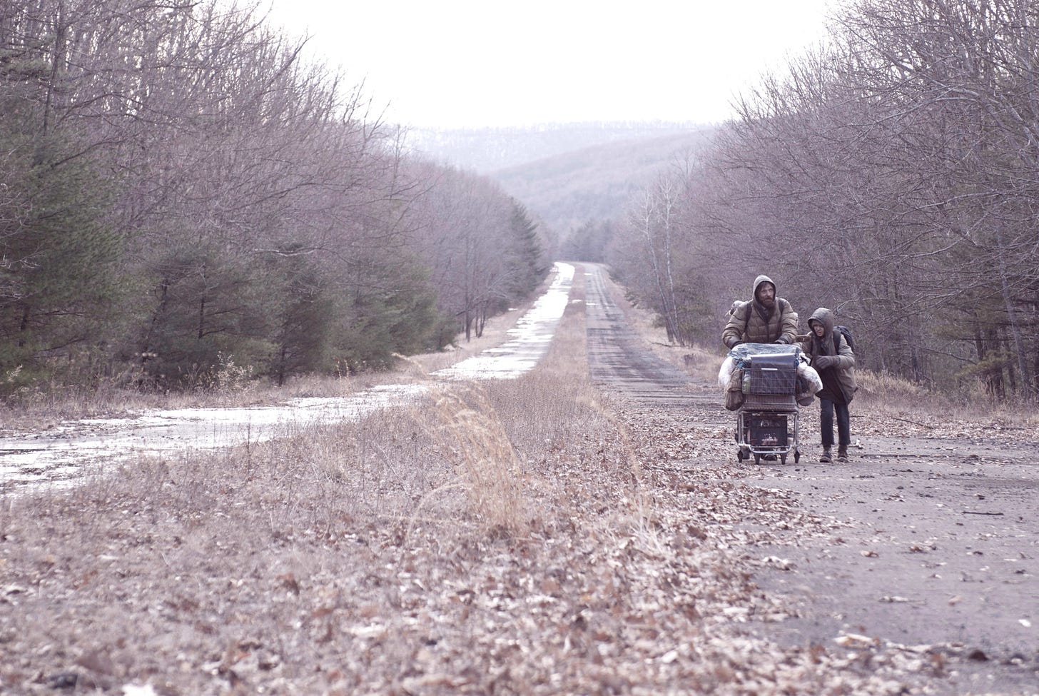 A man and his son push a shopping cart of stuff through a wasteland