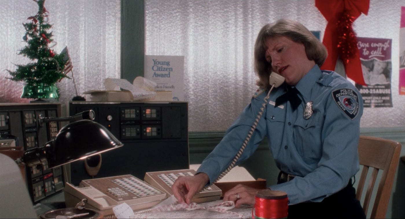 A police woman sitting at her desk, wrapping presents while talking on a chorded phone which she holds between her shoulder and her ear. On the shelf behind her is a decorative Christmas tree and an award that reads "Young Citizen Award"