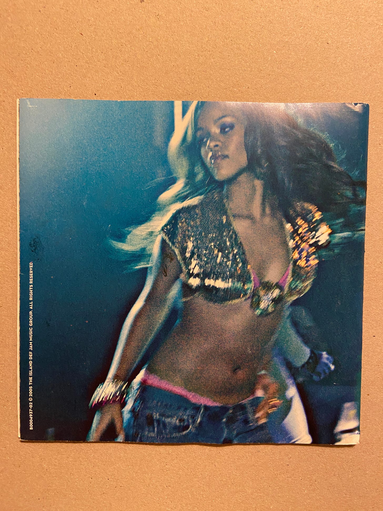 a still image from Rihanna's video for "Pon de Replay" as featured in her album for Music of the Sun (2005). she glamorously looks to the left while wearing a gold sparkle crop top and blue jeans with pink/red underwear peeking through.