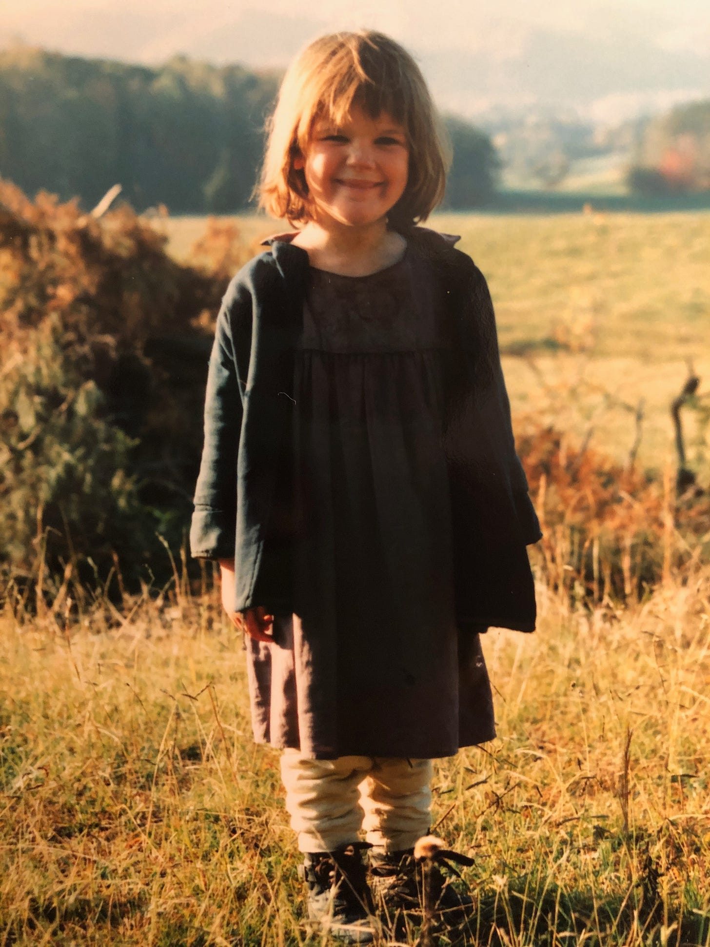 Katharine Duckett, pictured here at 6 or 7 years old, stands in a field on a sunny day, wearing a costume for the period drama "Christy," set in 1912 in East Tennessee. She is white, with brown bobbed hair, and wearing a brown dress, dark blue coat, beige leggings, and brown boots.