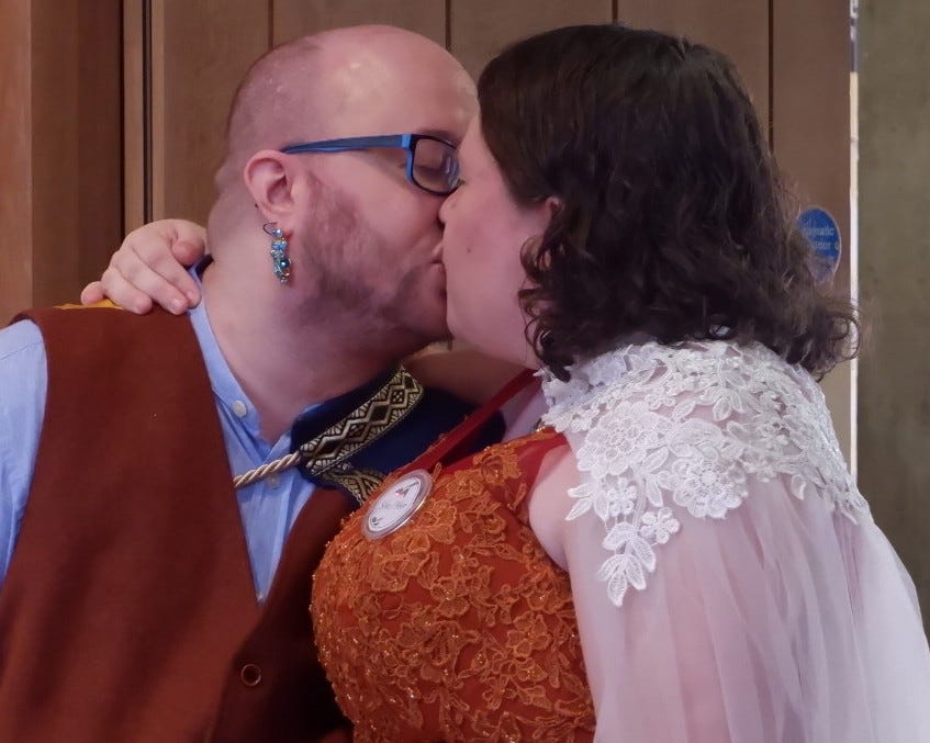 A beardgender person, with a shaved head and close-trimmed facial hair, wearing black & blue glasses, a dangly blue earring, a burnt orange waistcoat, light blue shirt, and royal blue cape, kisses a woman with brown hair, curly at the bottom, who wears a burnt orange dress with embroidery, and a white lacy train.
