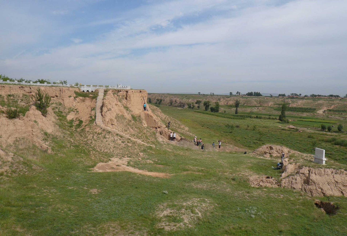 A sharp cliff face composed of tan sediment, with archaeologists working on a dig and surrounding green grassy valley A sharp cliff face composed of tan sediment, with archaeologists working on a dig and surrounding green grassy valley