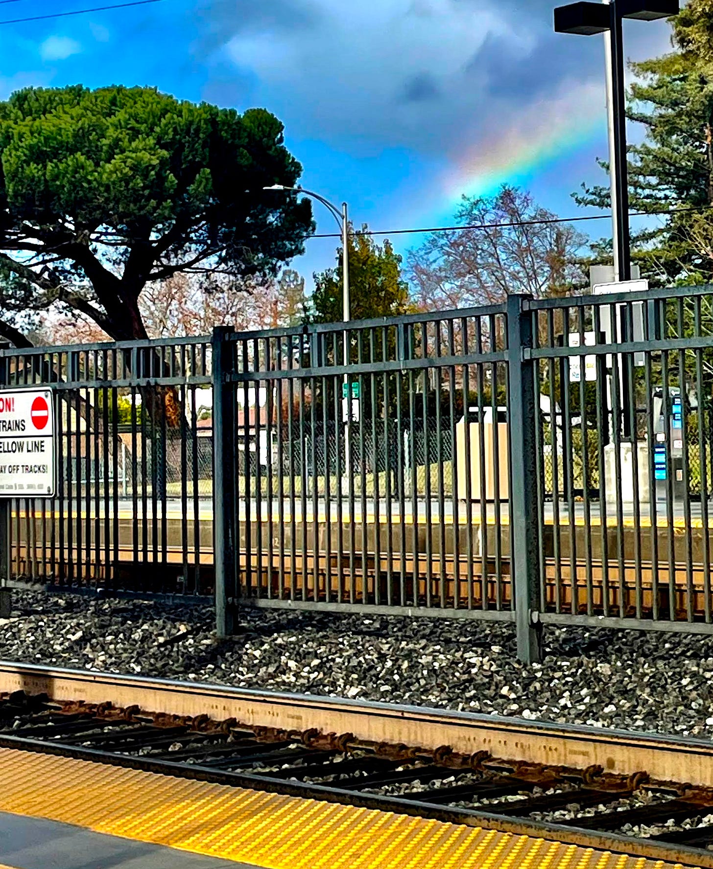 foreground: train stop with black bar fence separating the tracks. background: rainbow