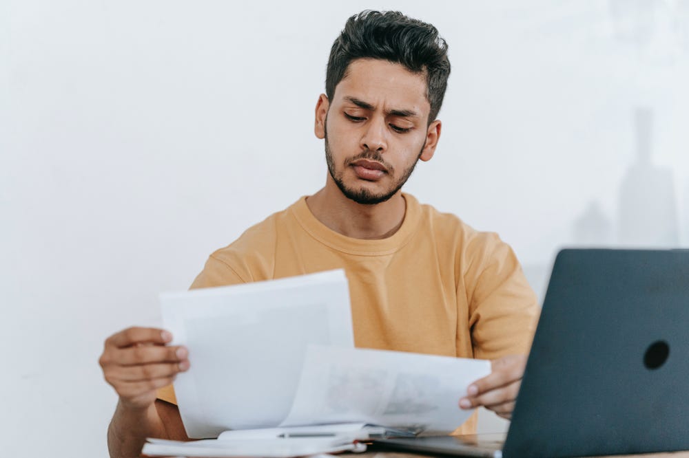 A young man with a neatly trimmed beard and mustache sitting in front of a laptop and looking at paperwork.