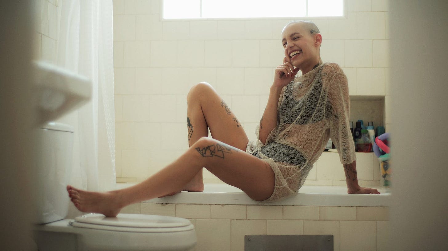 A woman sitting in the bathroom, on the lip on her bathtub, with one leg on the toilet, smiling and touching her face.