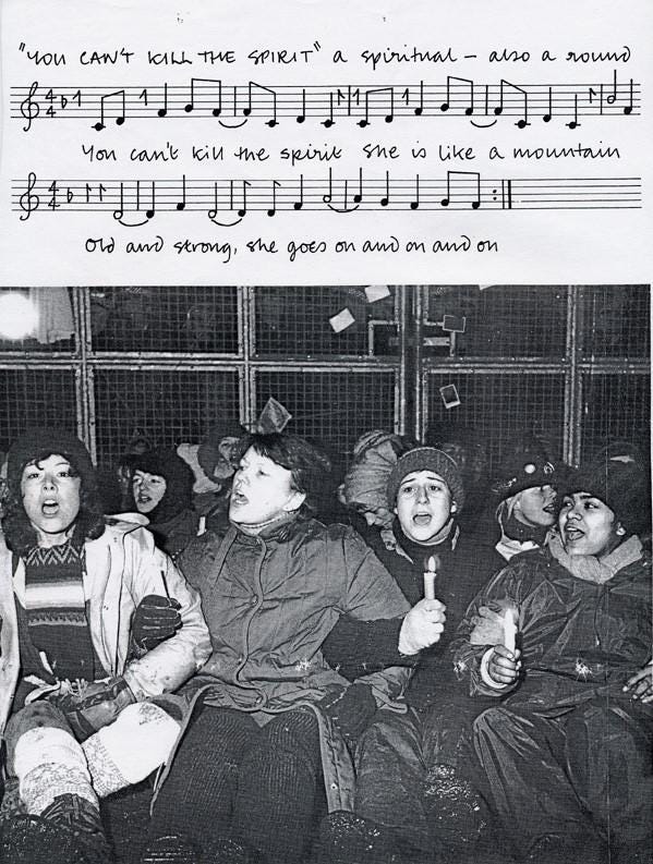 Photograph of a group of women sitting outside the gates to Greenham Common, holding candles and singing. Above is a fragment from a protest song. 
