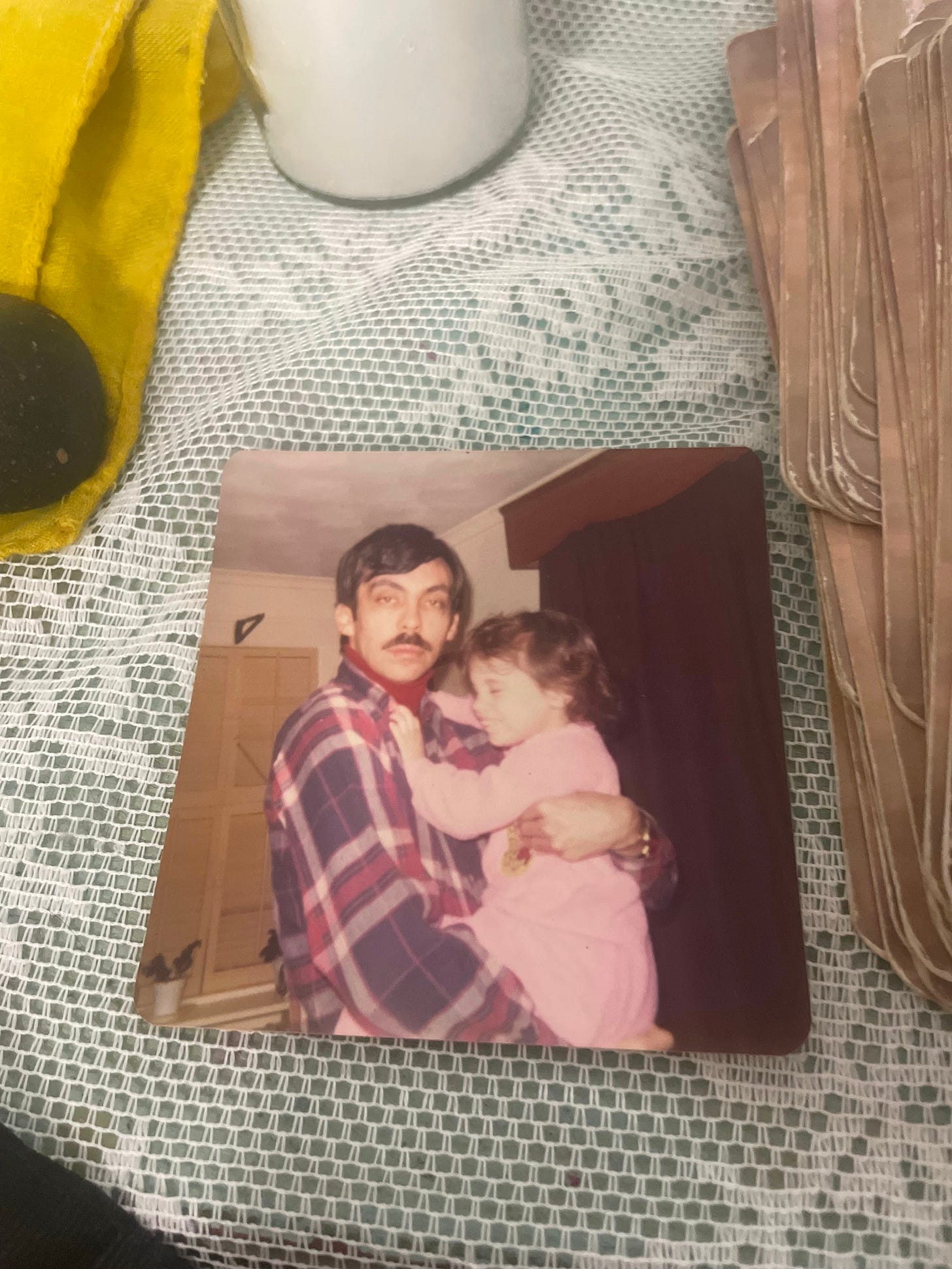 1970s square photo of Leah's dad, a light brown skinned man with thick black hair and a thick black moustache, wearing a plaid shirt, looking furious, diassociated and terrified, holding Leah, a light skinned three year old laughing and kicking in a pink onesie, the photo lies on a white lace tablecloth.