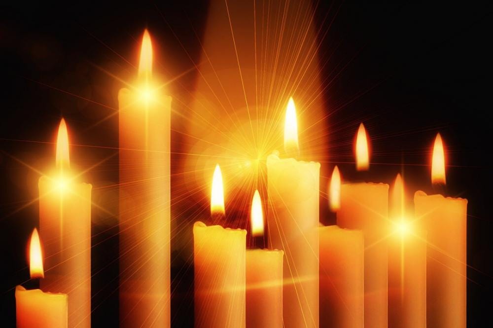 A cluster of white candles of different heights stand lit against a dark background.