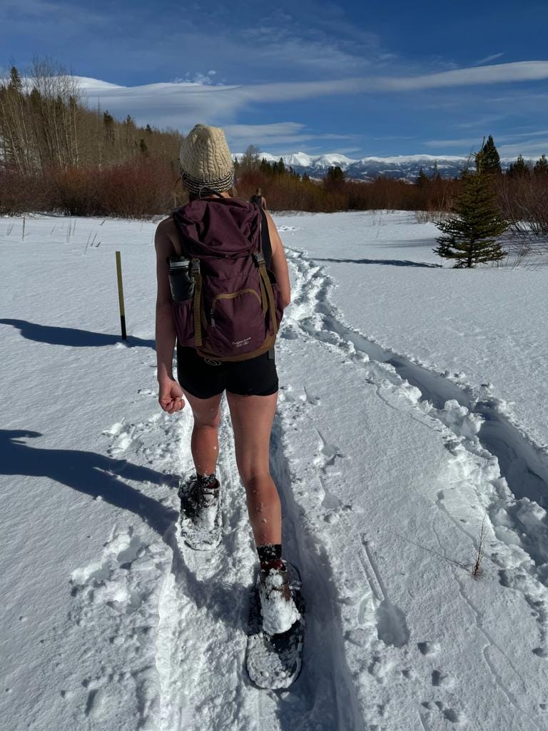 Lisa walking away from camera in snow, wearing snowshoes and athletic shorts, mountains in background