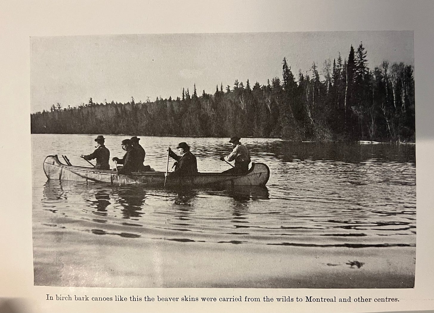 Black and white photo from an old book of five white men in hats and sport jackets, once of them smoking a pipe, paddling a birch bark canoe