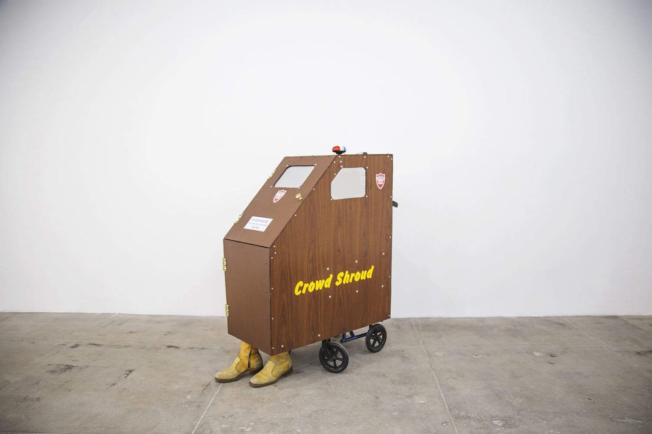 A wheelchair is sitting underneath a wooden car-shaped box, that reads Crowd Shroud on the side in yellow text. There are small cutouts for the driver to see out of, and there are yellow boots in the front, which indicates someone is sitting in the wheelchair. A wheelchair is sitting underneath a wooden car-shaped box, that reads Crowd Shroud on the side in yellow text. There are small cutouts for the driver to see out of, and there are yellow boots in the front, which indicates someone is sitting in the wheelchair.