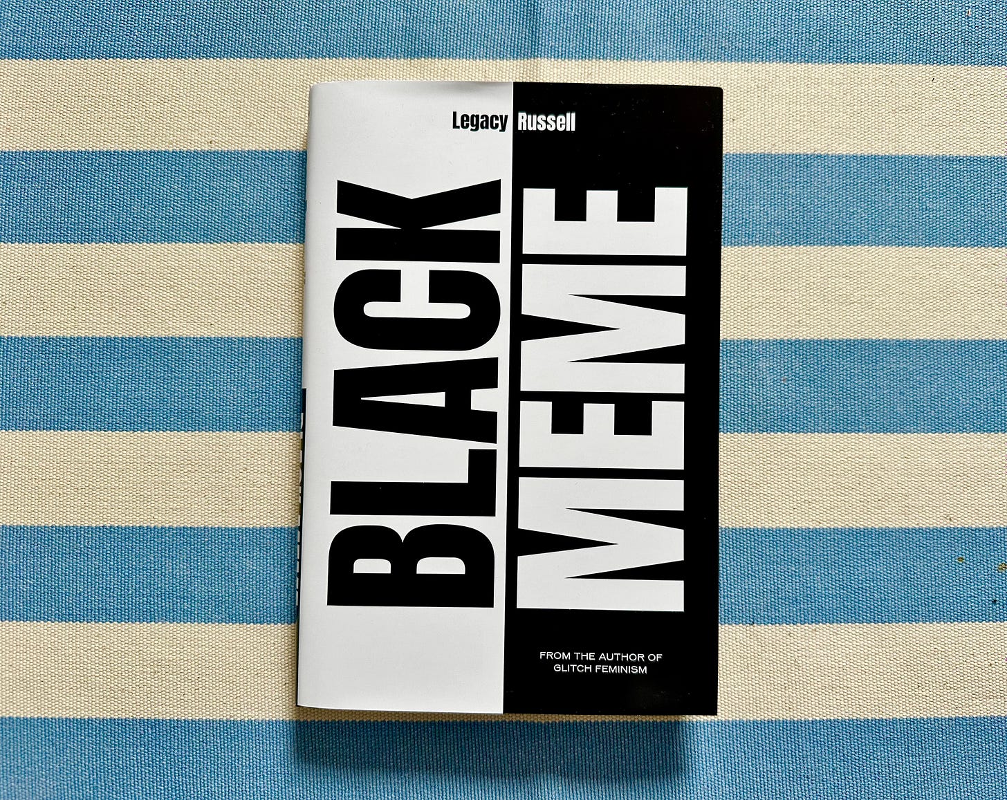 Picture of a book titled "Black Meme", by Legacy, on a textured cotton tablecloth with thick off-white and light blue stripes. The cover is a bold black and white design with the words appearing vertically — BLACK in black text on white background, MEME in whit text on a black background.