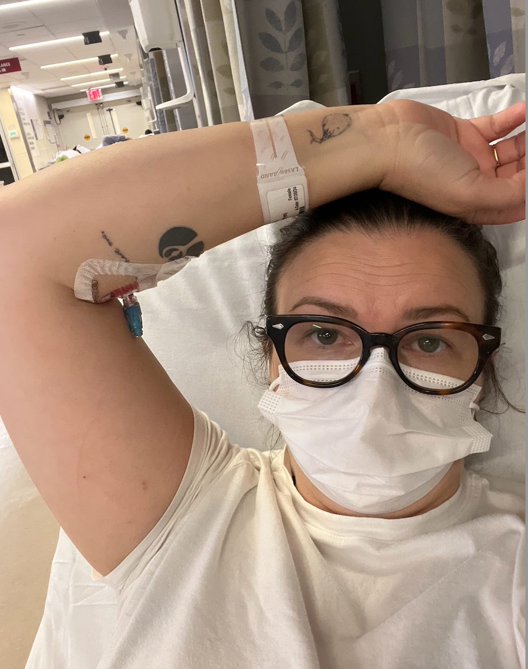 A selfie of Amber lying in a hospital bed. She wears black glasses, a white face mask, a hospital gown, and a hospital patient ID bracelet. She holds one arm up to show her IV.