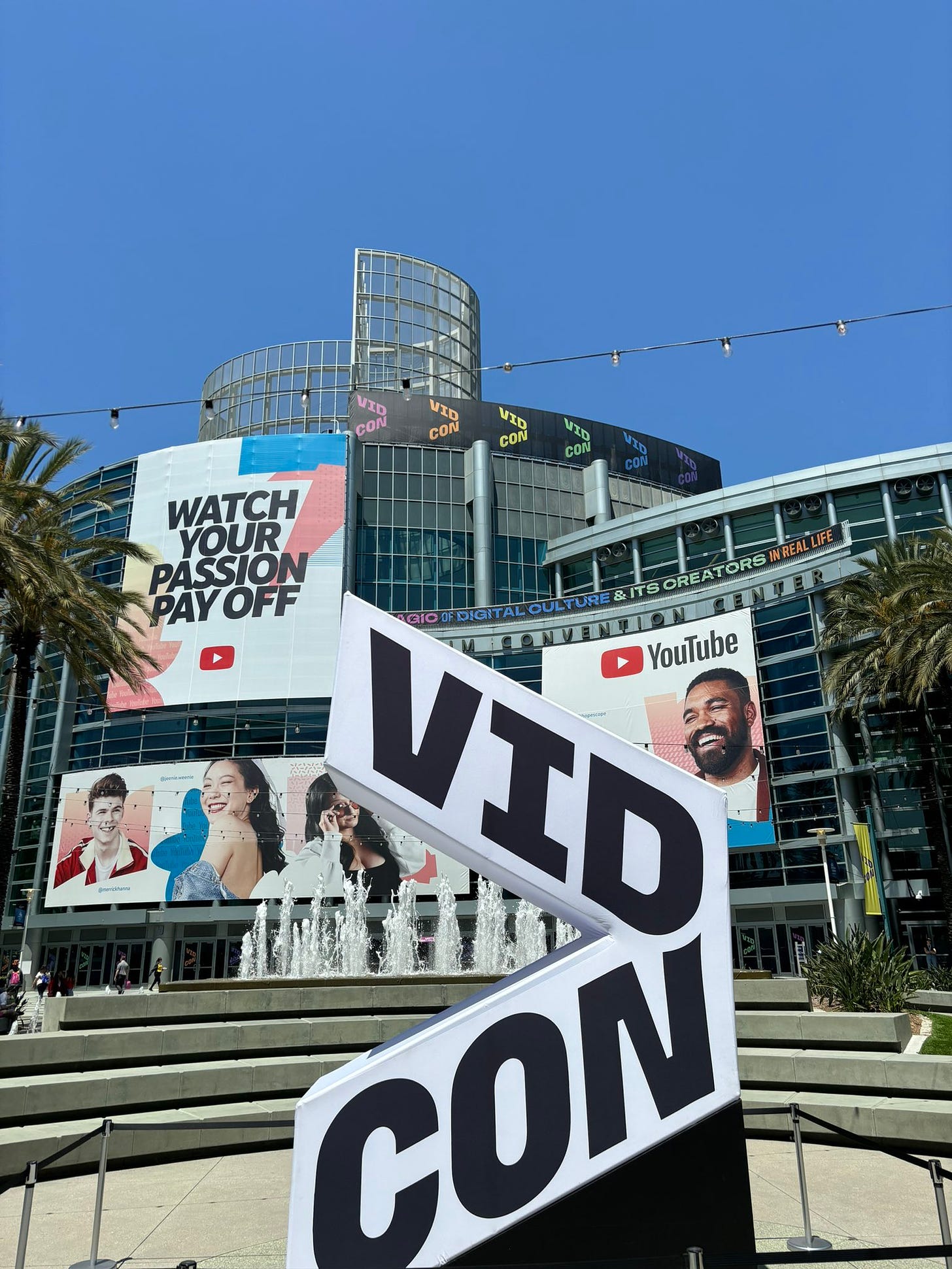 Screenshot of a VidCon conference image. In the foreground is a large sign that says VidCon. The Anaheim Convention Center is in the background. A banner on the building says: Watch your passion pay off