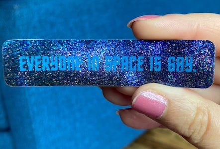 a hand holding a sparkly blue "everyone in space is gay" sticker
