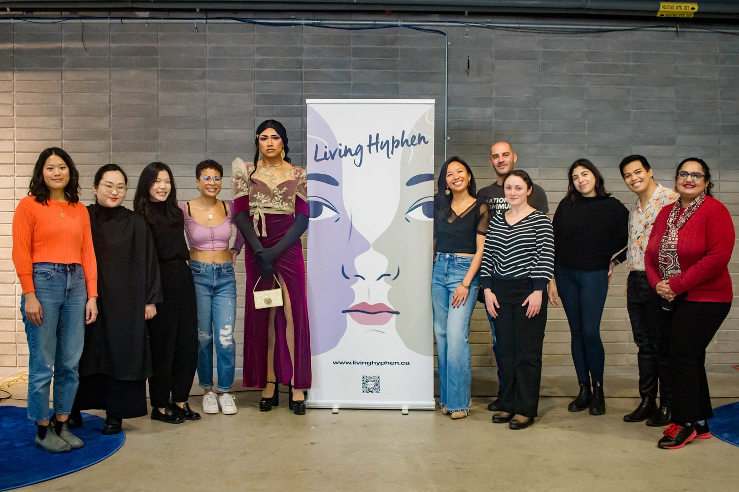 Group of Living Hyphen performers standing and smiling next to a giant banner with the LH logo. 
