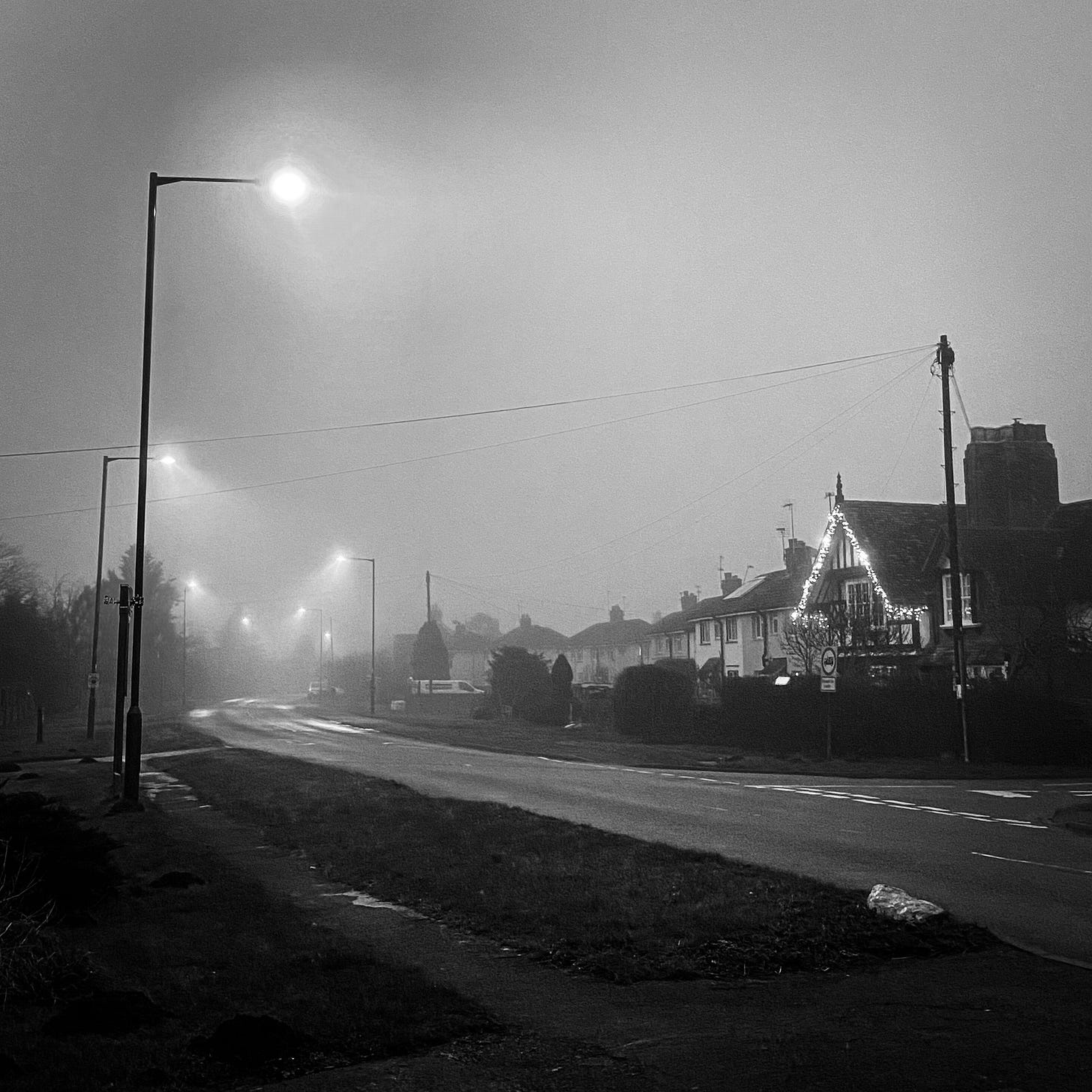 A black and white misty village scene, its curving road illuminated beneath L E D street lamps.