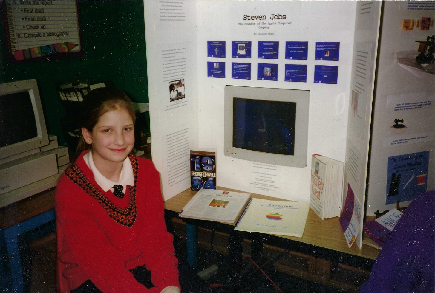The author as a child wearing a tie and red sweater sitting in front of a posterboard that reads "Steven Jobs: The Found of the Apple Computer Company." An Apple monitor and several books are on display.