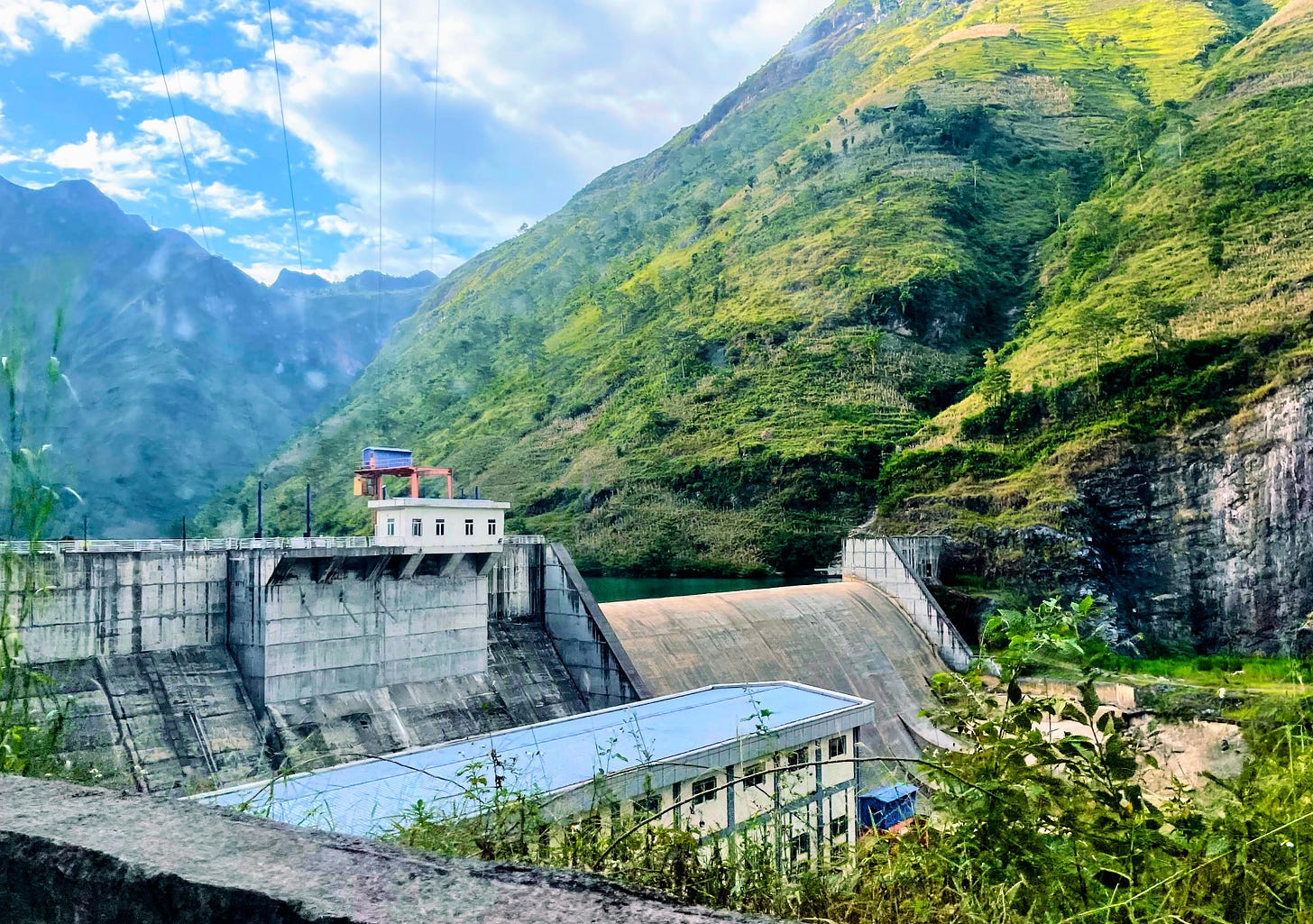 A large concrete dam sits in a deep, green river valley, with dramatic peaks in the backdrop under a partly-cloudy sky. A large concrete dam sits in a deep, green river valley, with dramatic peaks in the backdrop under a partly-cloudy sky.
