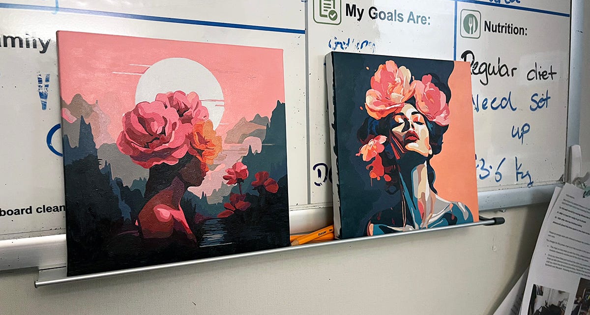 Two completed paint-by-number canvases propped up against a hospital whiteboard. Both feature beautiful women with enormous pink and orange flowers in their hair.