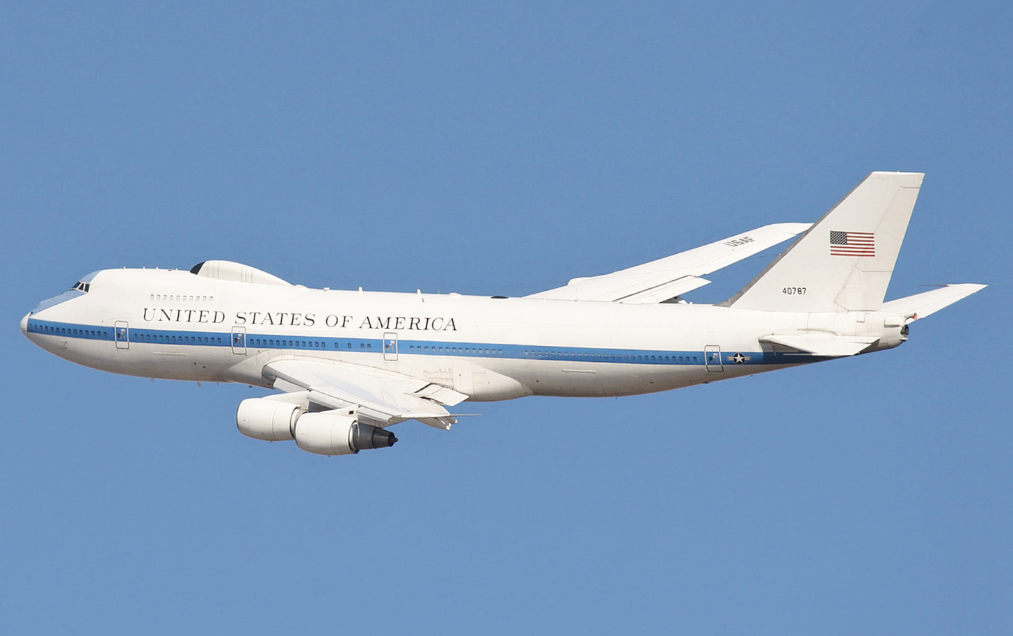 White jet with blue cheatline in-flight over land, flying left. The aircraft features a distinctive bump above the usual 747's trademark nose profile